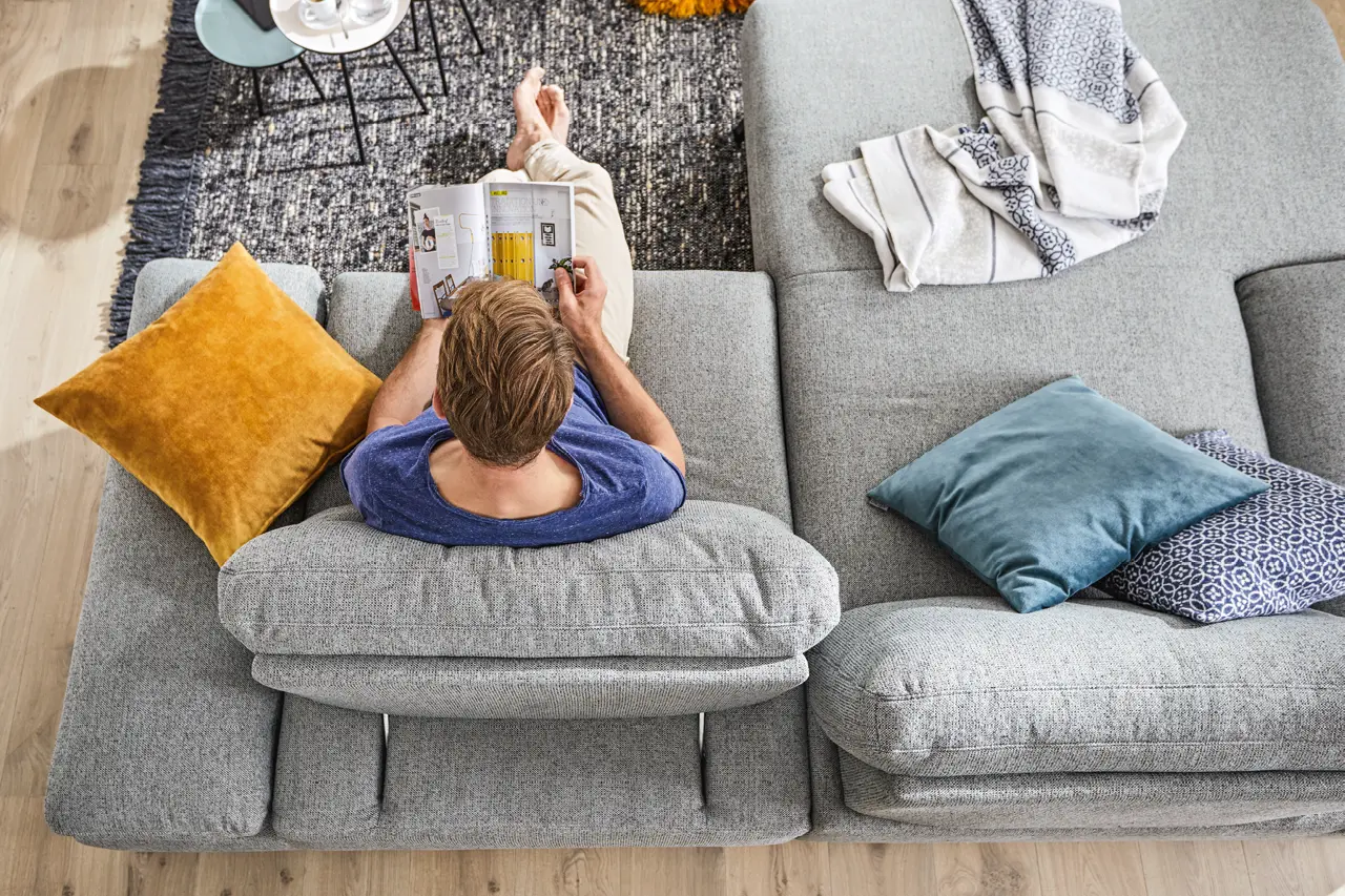 Mann entspannt auf einer grauen Wohnlandschaft und liest ein Magazin, gemütliches Wohnzimmer mit Dekokissen, Decke und Teppich, modernes Sofa mit hohem Sitzkomfort und wohnlicher Atmosphäre